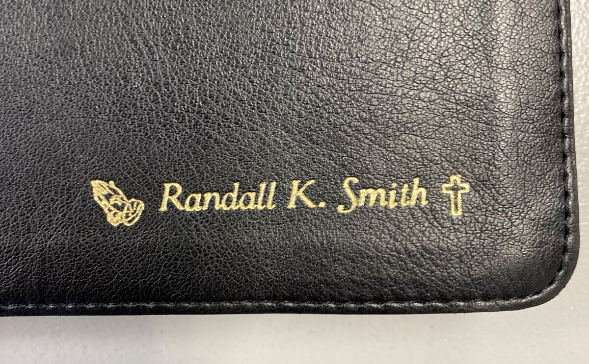 Gold name imprint between praying hands and cross symbols on a black Bible